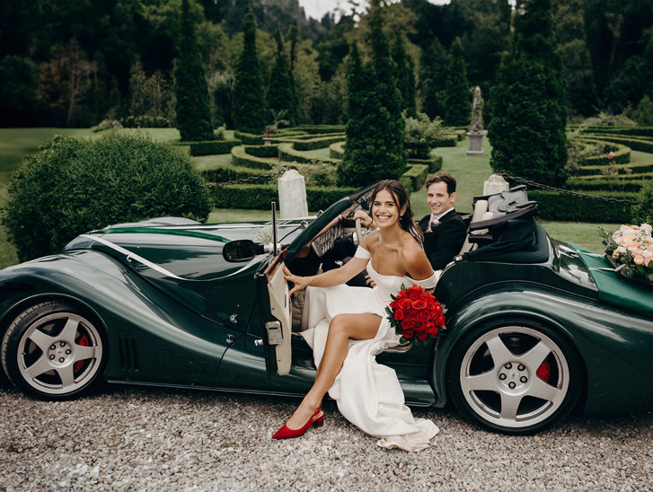 Bride and groom in sports car, bride is wearing red shoes and holding a bouquet of red roses